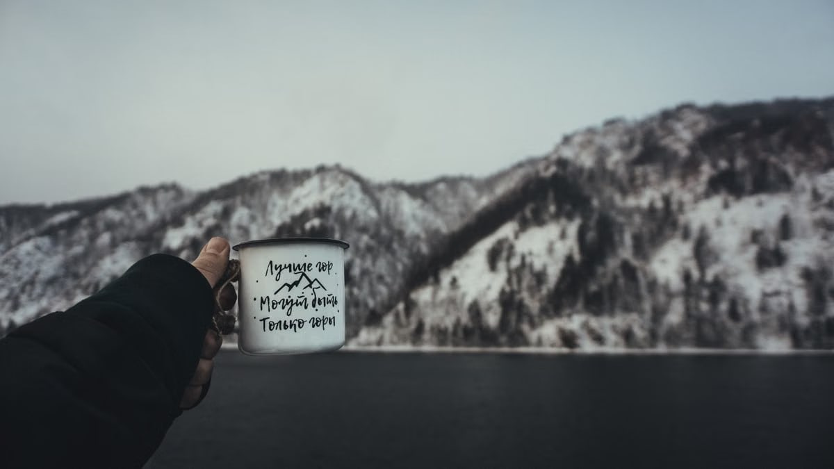 A scenic winter view of a hand holding a cup against snowy mountains and a lake.